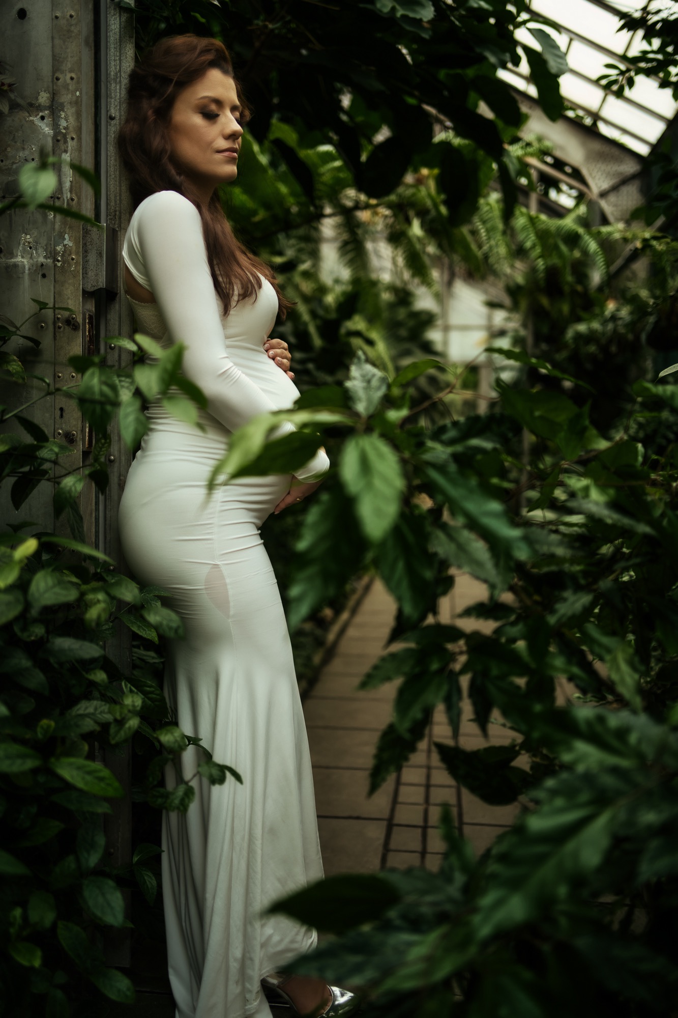 Kobieta w obcisłej białej sukni, opierająca się o metalowe drzwi w tropikalnym ogrodzie, z rękami na brzuchu