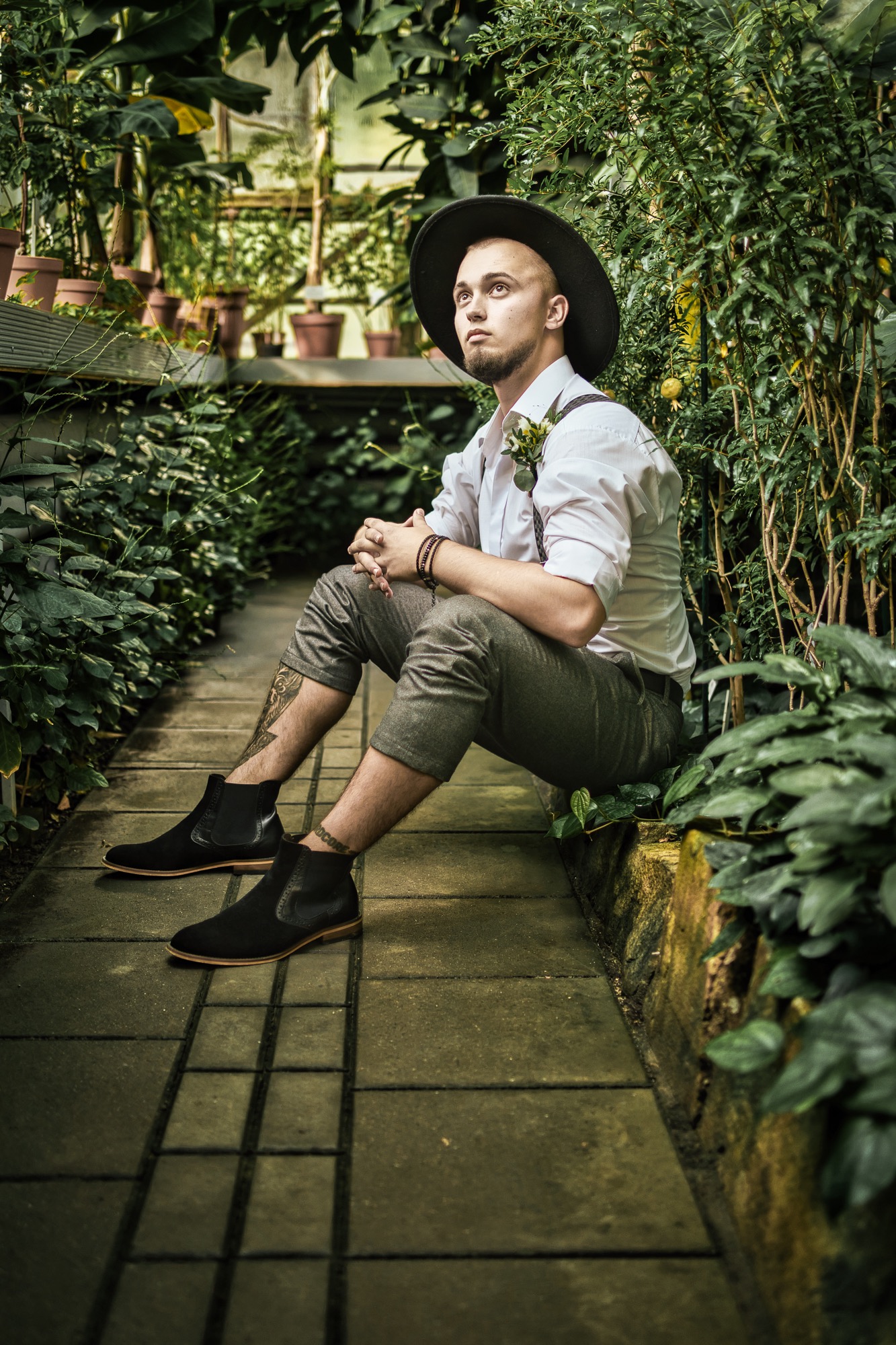 Młody mężczyzna w kapeluszu i szelkach siedzi w szklarni, oparty o ścianę porośniętą roślinami, z nogami skrzyżowanymi i dłońmi złożonymi na kolanach, patrząc w górę.