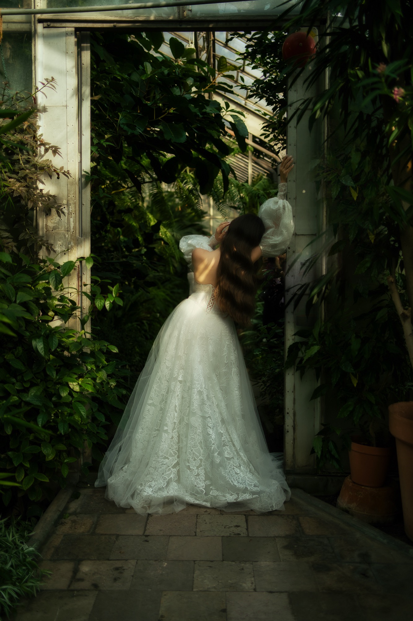 Kobieta w koronkowej sukni ślubnej z odkrytymi plecami, opierająca się o przeszklone drzwi w otoczeniu bujnej zieleni w szklarni.