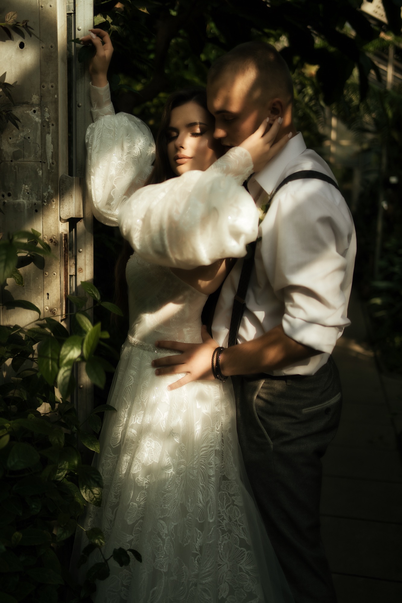 Para młoda w objęciach przed metalową konstrukcją porośniętą zielenią, panna młoda w sukni z bufiastymi rękawami, pan młody w szelkach, miękkie światło.