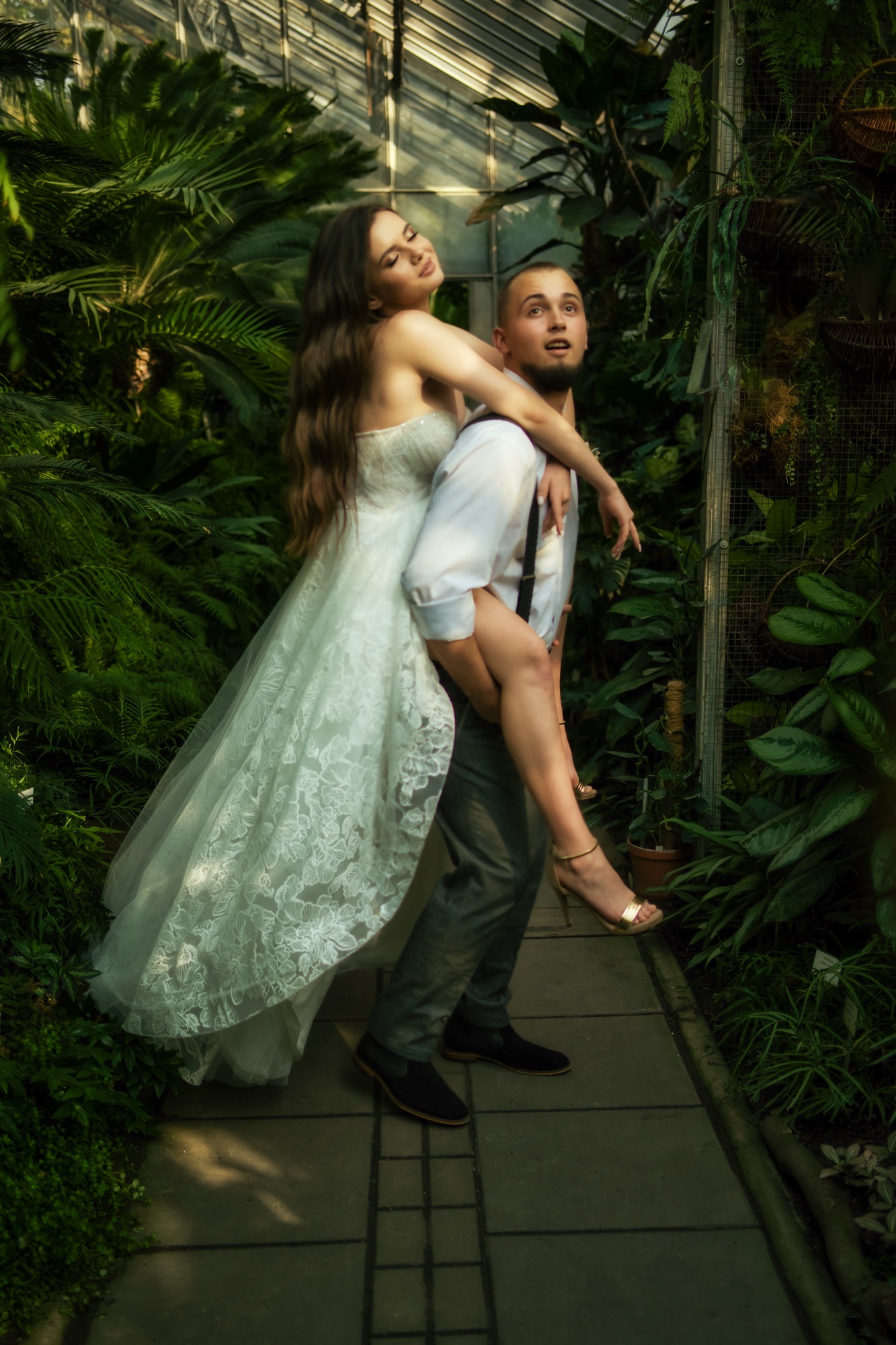 Para w tropikalnej szklarni: mężczyzna niesie kobietę w białej sukni ślubnej, ona uśmiechnięta obejmuje go za szyję, tło stanowią bujne rośliny.