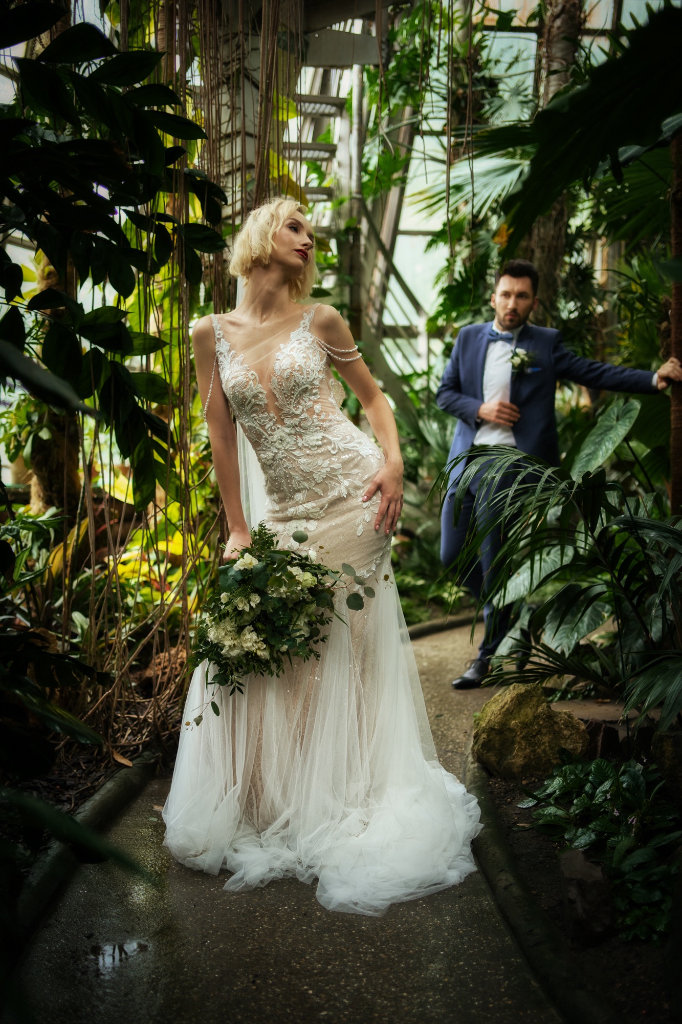 Panna młoda w koronkowej sukni ślubnej z bukietem, pozująca w szklarni, obok stoi pan młody w niebieskim garniturze.