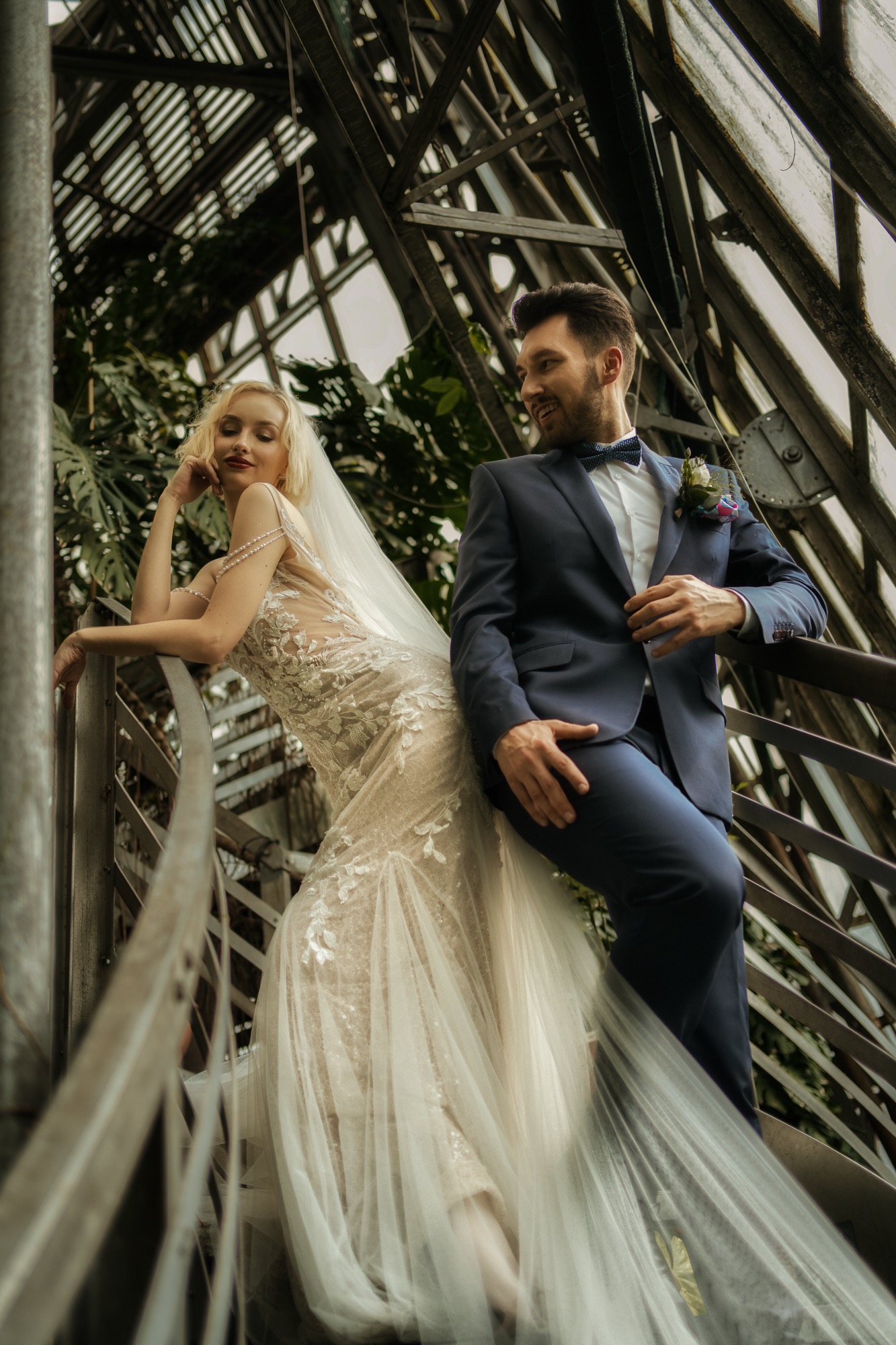 Para młoda pozuje na metalowych schodach w szklarni; panna młoda w koronkowej sukni z welonem opiera się o poręcz, pan młody w granatowym garniturze z muszką patrzy na nią z uśmiechem.