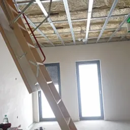 Montaż drewnianych schod&oacute;w strychowych z metalowymi elementami; widoczne profile sufitowe, ocieplenie natryskowe, okna w tle oraz narzędzia i materiały budowlane w pomieszczeniu w stanie surowym.