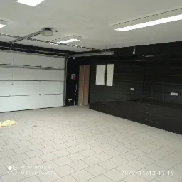 Wyłożona jasnymi płytkami podłoga w pomieszczeniu z czarną ścianą z płytek i białą bramą garażową, widoczne puste puszki elektryczne na ścianie.