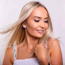 Portret uśmiechniętej, blondwłosej kobiety z delikatnym makijażem, perłowymi kolczykami i złotym łańcuszkiem na białym tle.