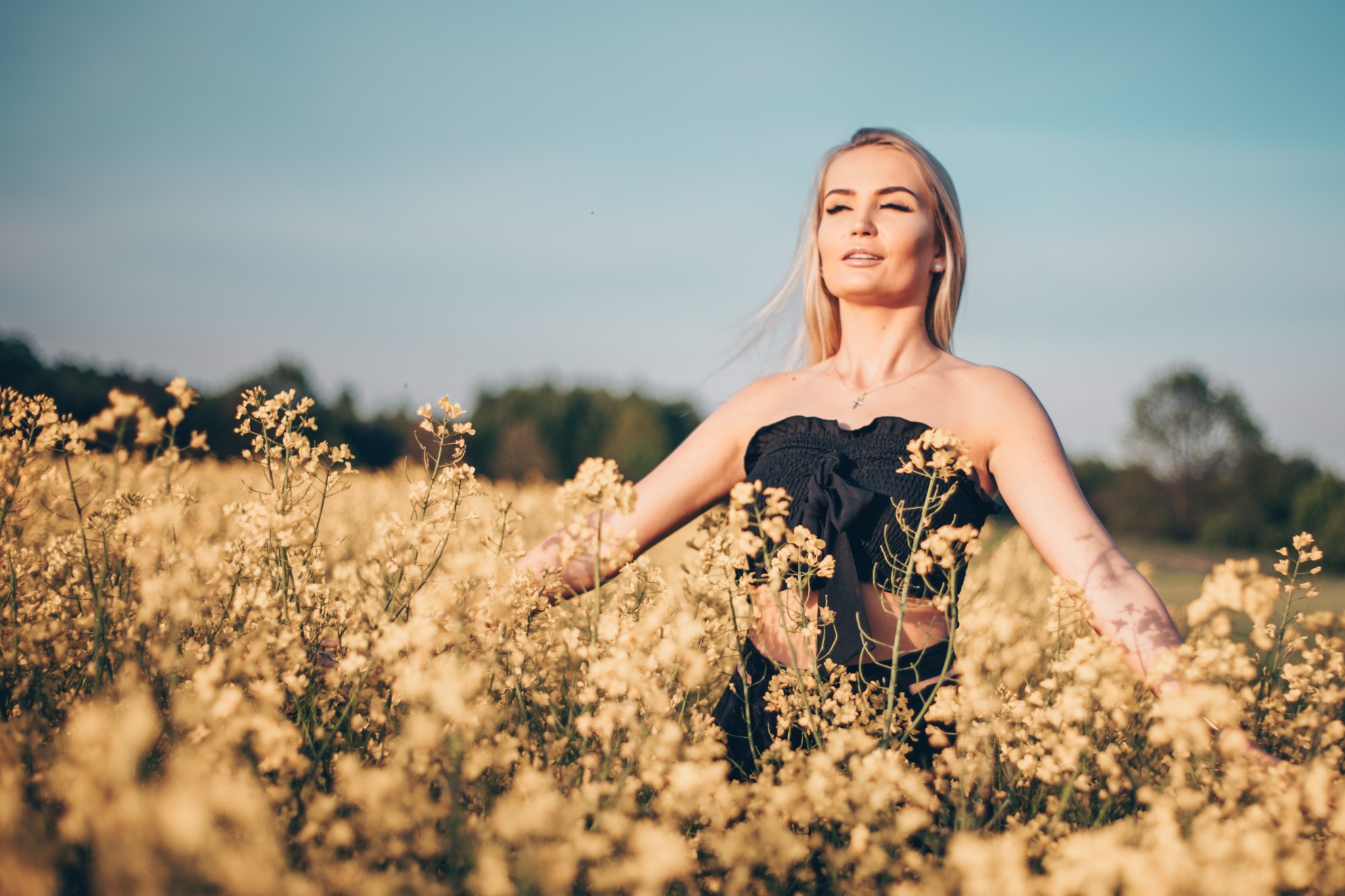 Kobieta w czarnej sukience stoi w polu kwitnącego rzepaku, z rozłożonymi ramionami, spoglądając w górę na niebo.