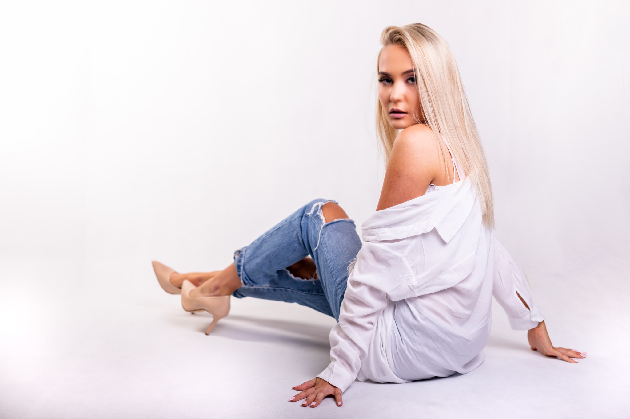 Blondwłosa kobieta w podartej jeansowej spodniach i białej koszuli pozuje siedząc na białym tle, spoglądając przez ramię.
