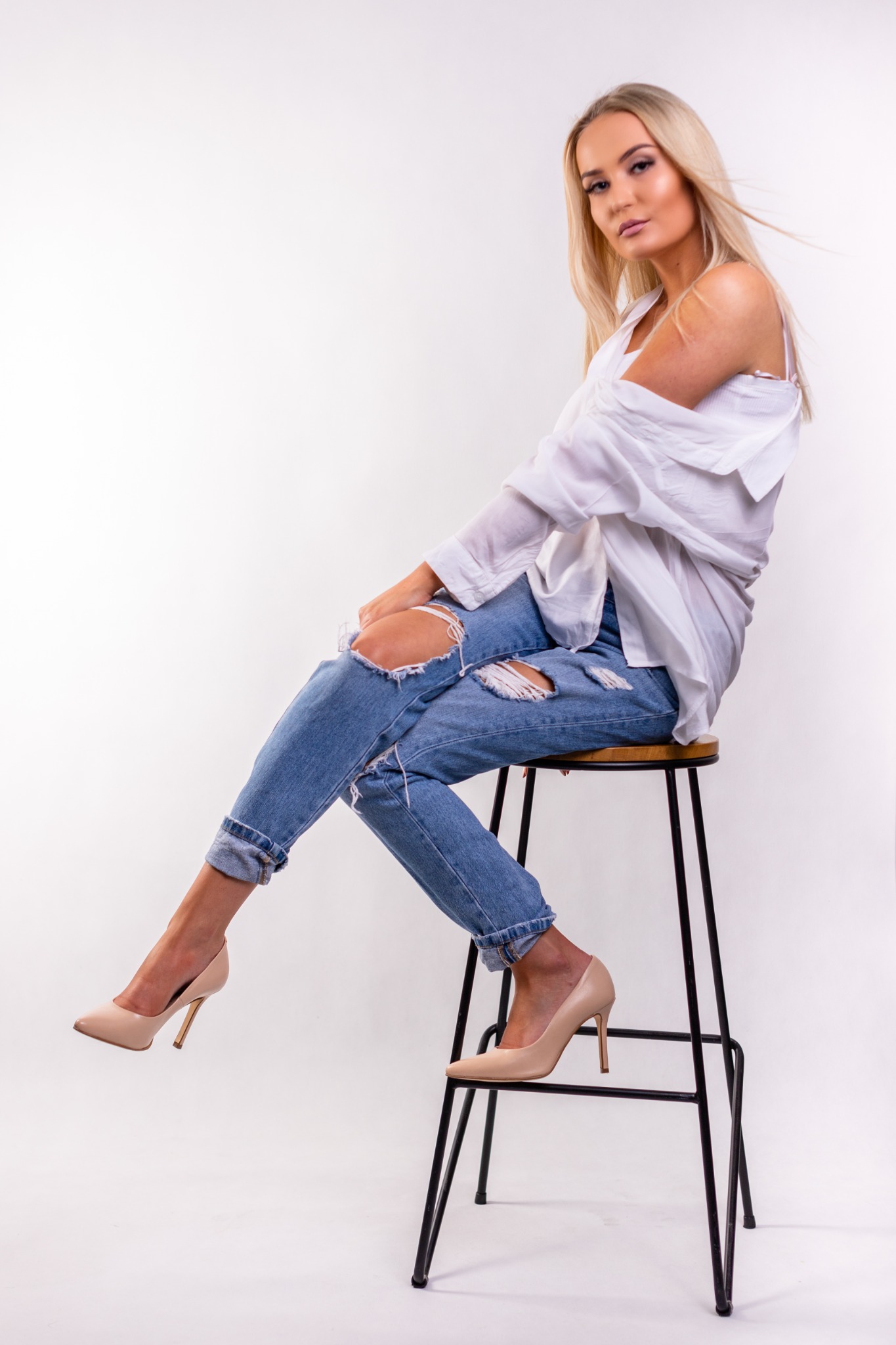Blondwłosa kobieta w rozpiętej białej koszuli i podartych dżinsach siedzi na wysokim stołku, prezentując beżowe szpilki na tle białego studia.