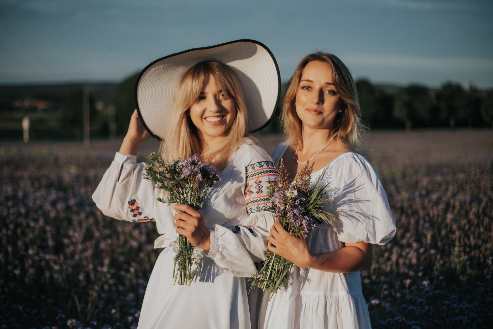 Dwie uśmiechnięte kobiety w białych sukienkach z haftem, trzymają bukiety polnych kwiatów na tle kwitnącego pola; jedna z nich ma na głowie duży kapelusz.