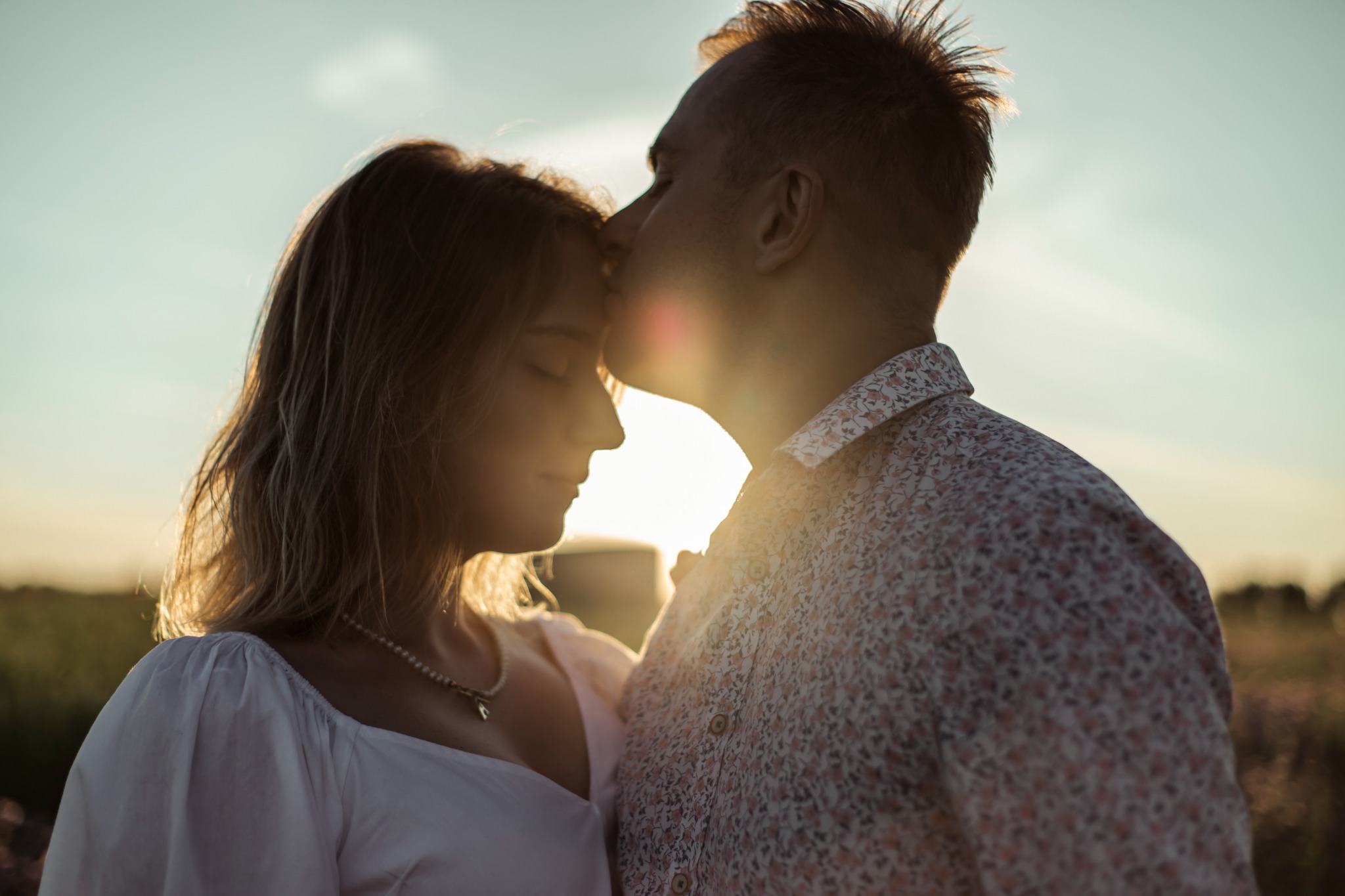 Silhouettes of a couple in love, man kissing woman's forehead against a golden sunset backdrop, she wears a delicate pearl necklace.