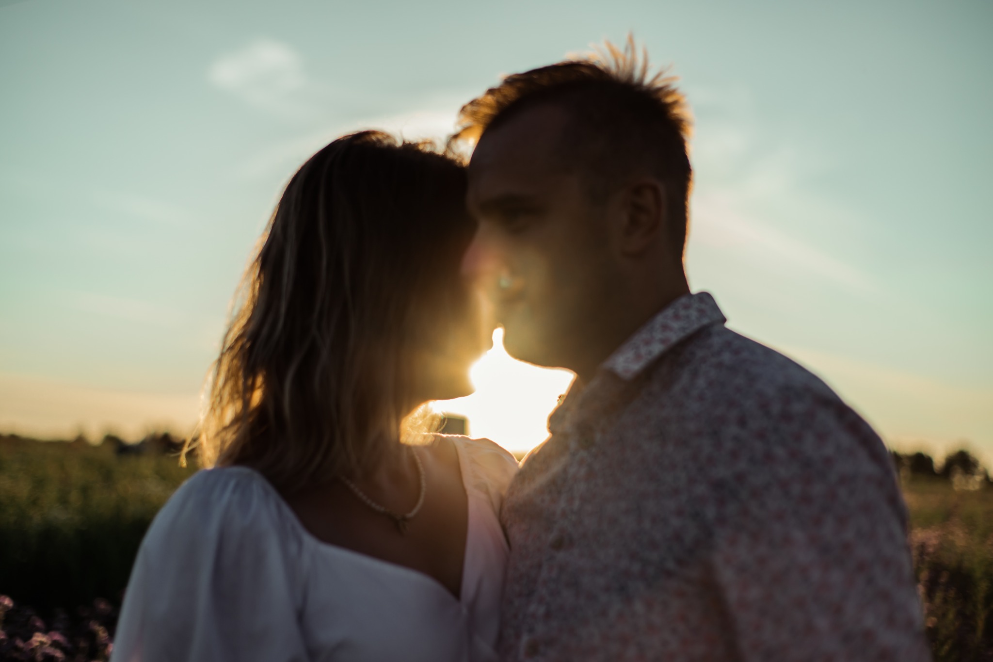 Para w kontraście ze słońcem, sylwetki zbliżone do pocałunku na tle łąki o zachodzie słońca, ona w białej sukience i perłowym naszyjniku, on w kwiecistej koszuli.