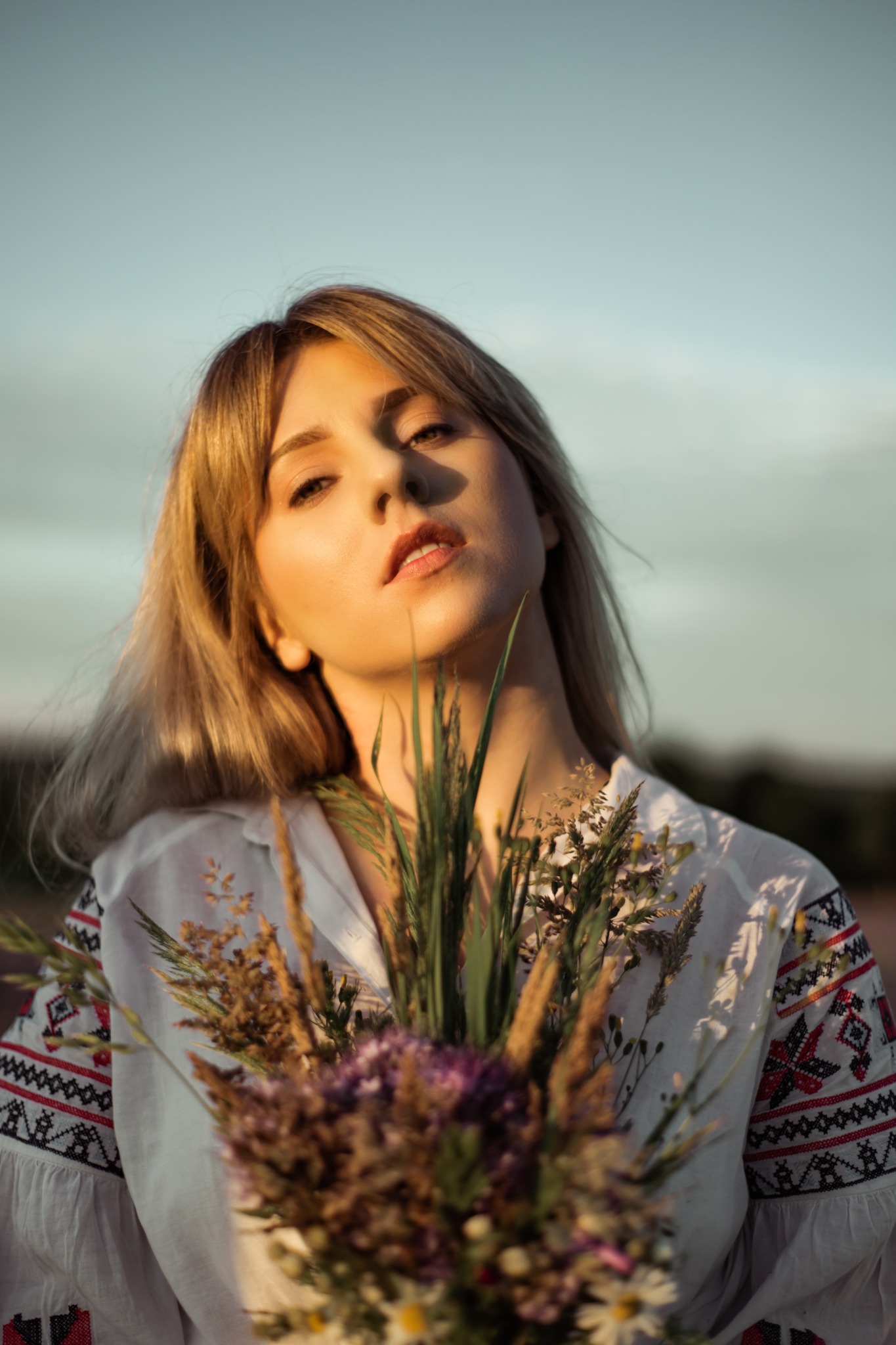 Portret kobiety w haftowanej bluzce, trzymającej bukiet polnych kwiatów w promieniach zachodzącego słońca.