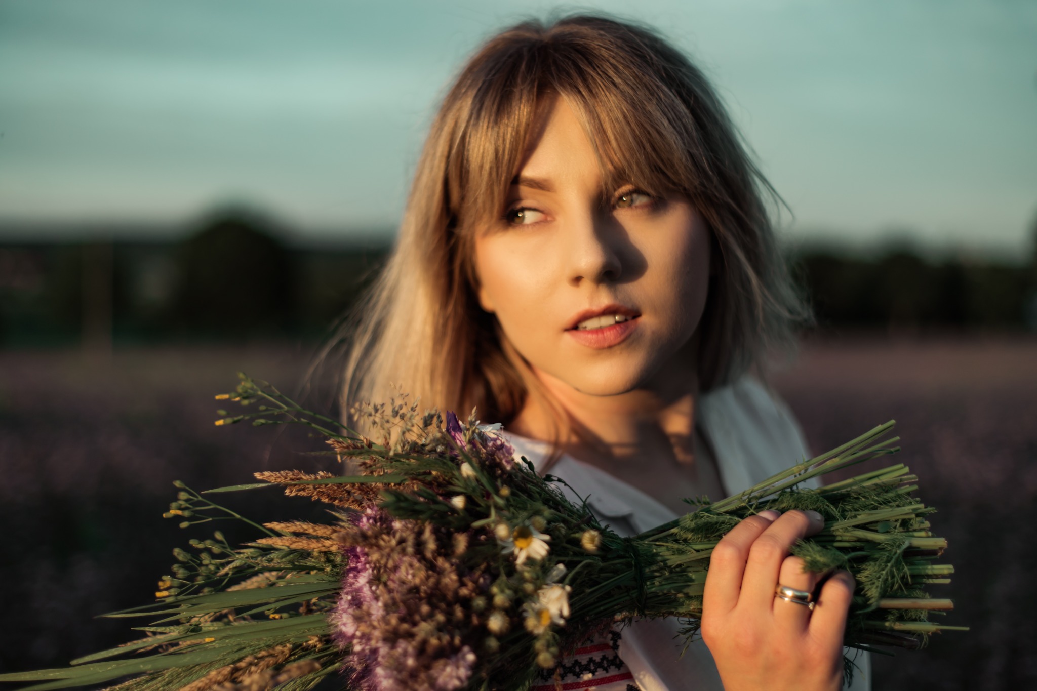 Portret młodej kobiety z blond włosami, trzymającej bukiet polnych kwiatów, w tle rozmyte pole i zachodzące słońce.