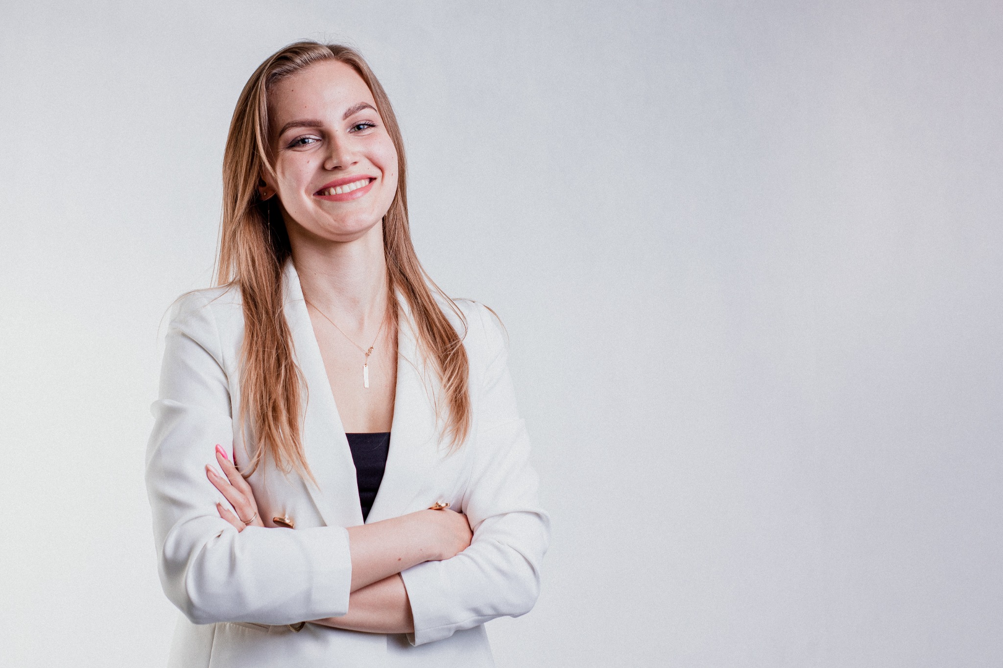 Portret uśmiechniętej kobiety w białym żakiecie na jasnym tle, z rękami skrzyżowanymi na piersiach i widoczną złotą biżuterią.