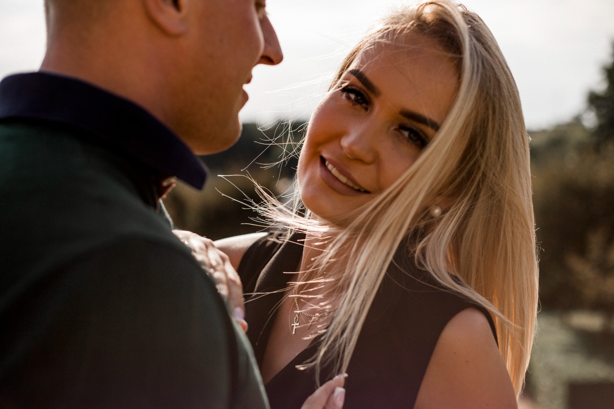 Uśmiechnięta blond kobieta z delikatnym makijażem, w czarnej sukience, obejmowana przez mężczyznę w ciemnozielonej koszuli polo, w tle rozmyta zieleń.