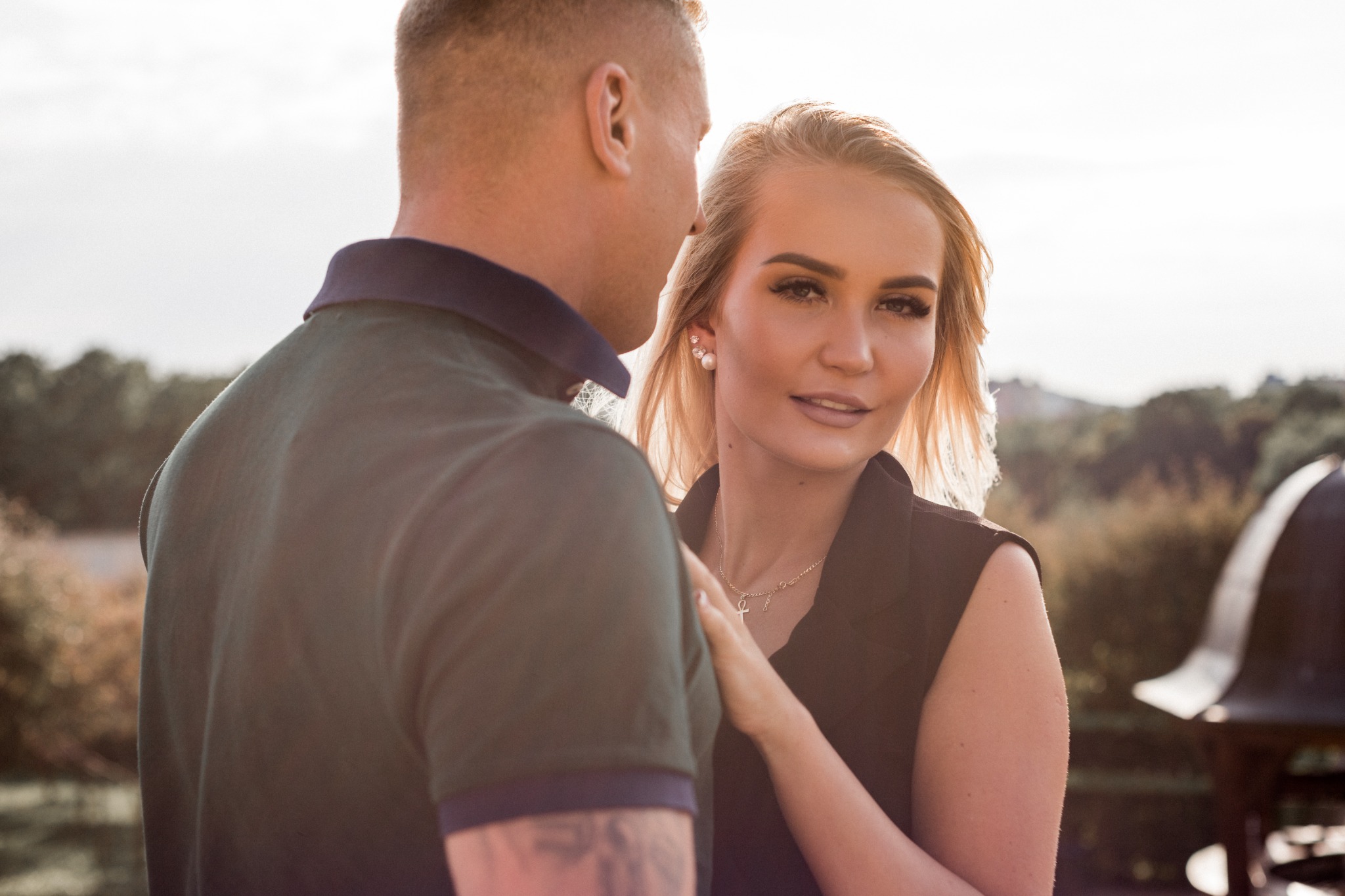 Zakochana para w delikatnym, ciepłym świetle. Kobieta z blond włosami i subtelnym makijażem spogląda w obiektyw, opierając dłoń na ramieniu mężczyzny w koszulce polo. W tle rozmyty krajobraz...