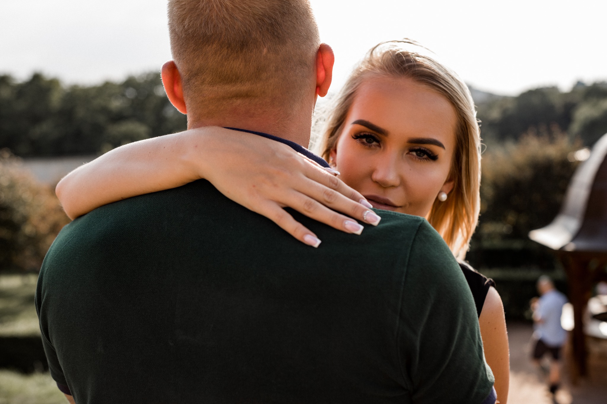 Blondwłosa kobieta obejmuje mężczyznę, patrząc w obiektyw, z widoczną dłonią z manicure francuskim i perłowym kolczykiem, na rozmytym tle zieleni i zarysu sylwetki.