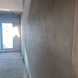 Świeżo nałożony tynk maszynowy na ścianie wewnętrznej, widoczne ślady po mocowaniu, w tle okno z niebieską ramą i wystającymi przewodami elektrycznymi przy podłodze.