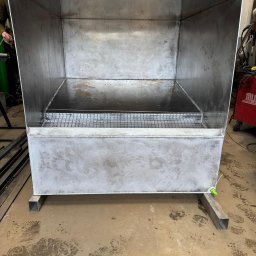 N&M Welding LTD - Surowa, metalowa konstrukcja przypominająca wannę spawalniczą na metalowych nogach, z kratką wewnątrz. Widoczne ślady pracy spawalniczej i niedoskonałości wykończenia.