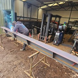 N&M Welding LTD - Spawanie metalowej belki H-kształtnej na kozłach w warsztacie, spawacz w masce, widoczny wózek widłowy i sprzęt spawalniczy. Podłoże ziemiste.