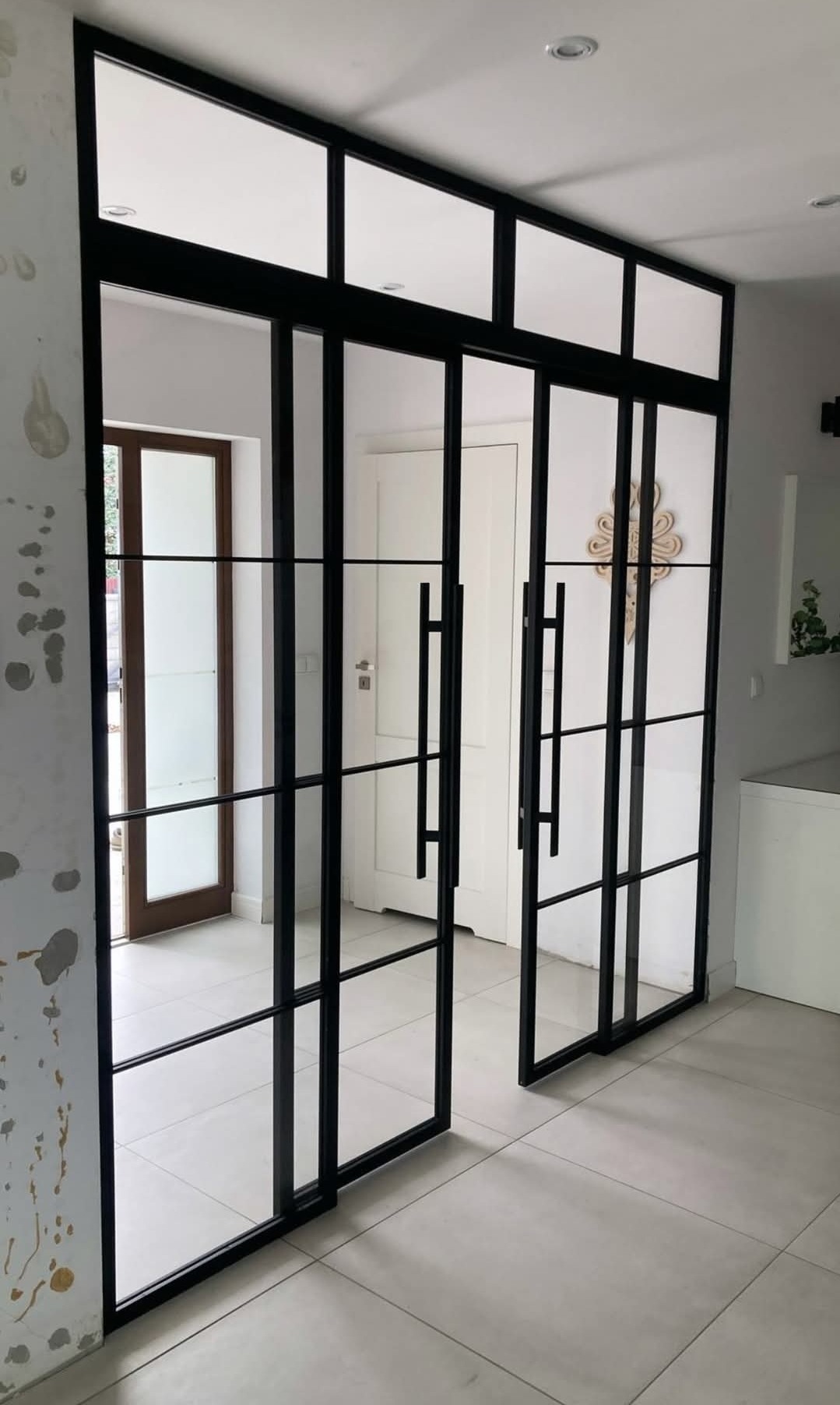 Nowoczesne, czarne drzwi metalowe z szybami w Chelmsford. Przeszklona konstrukcja z długimi uchwytami, widok na wnętrze domu i białe drzwi w tle.