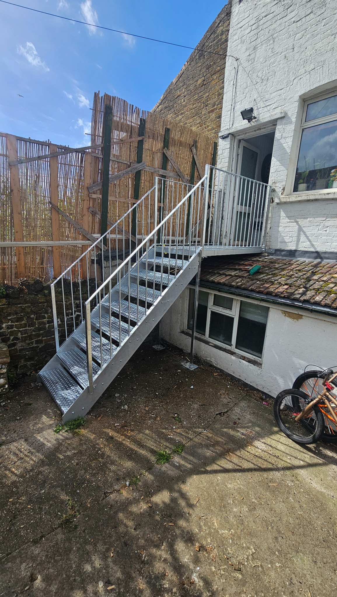 Zewnętrzne schody metalowe z balustradą, prowadzące do wejścia na piętrze. Widoczne elementy konstrukcyjne i otoczenie budynku. Metalowe schody ocynkowane.