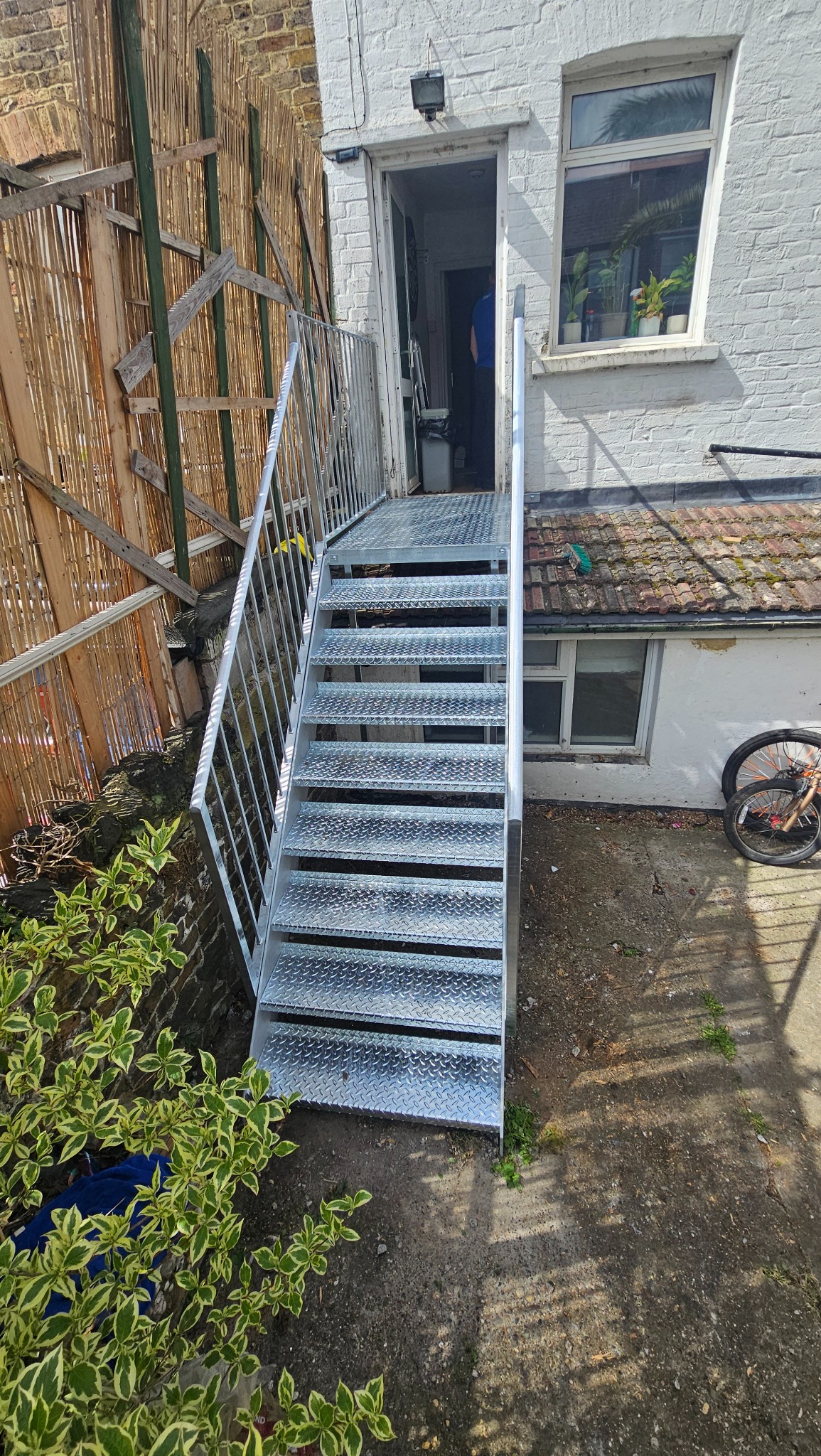 Zewnętrzne, metalowe schody z poręczą prowadzące do drzwi budynku. Schody wykonane z blachy ryflowanej. W tle fragment ściany budynku i elementu ogrodzenia.