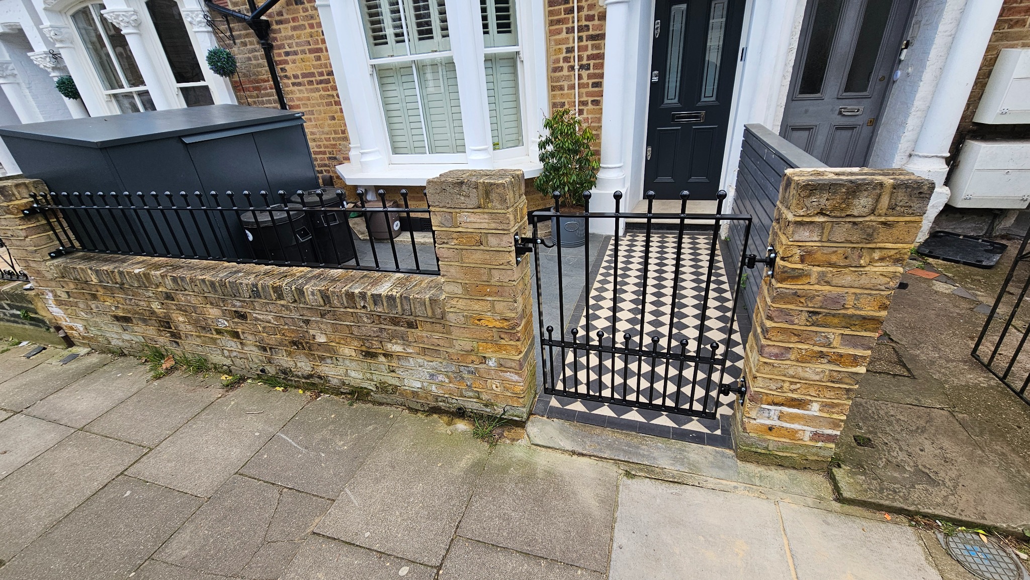 Czarne, metalowe ogrodzenie z bramką, zamontowane do ceglanego murku i słupków, z wzorem szachownicy na podłożu za bramką. Ujęcie z perspektywy chodnika.