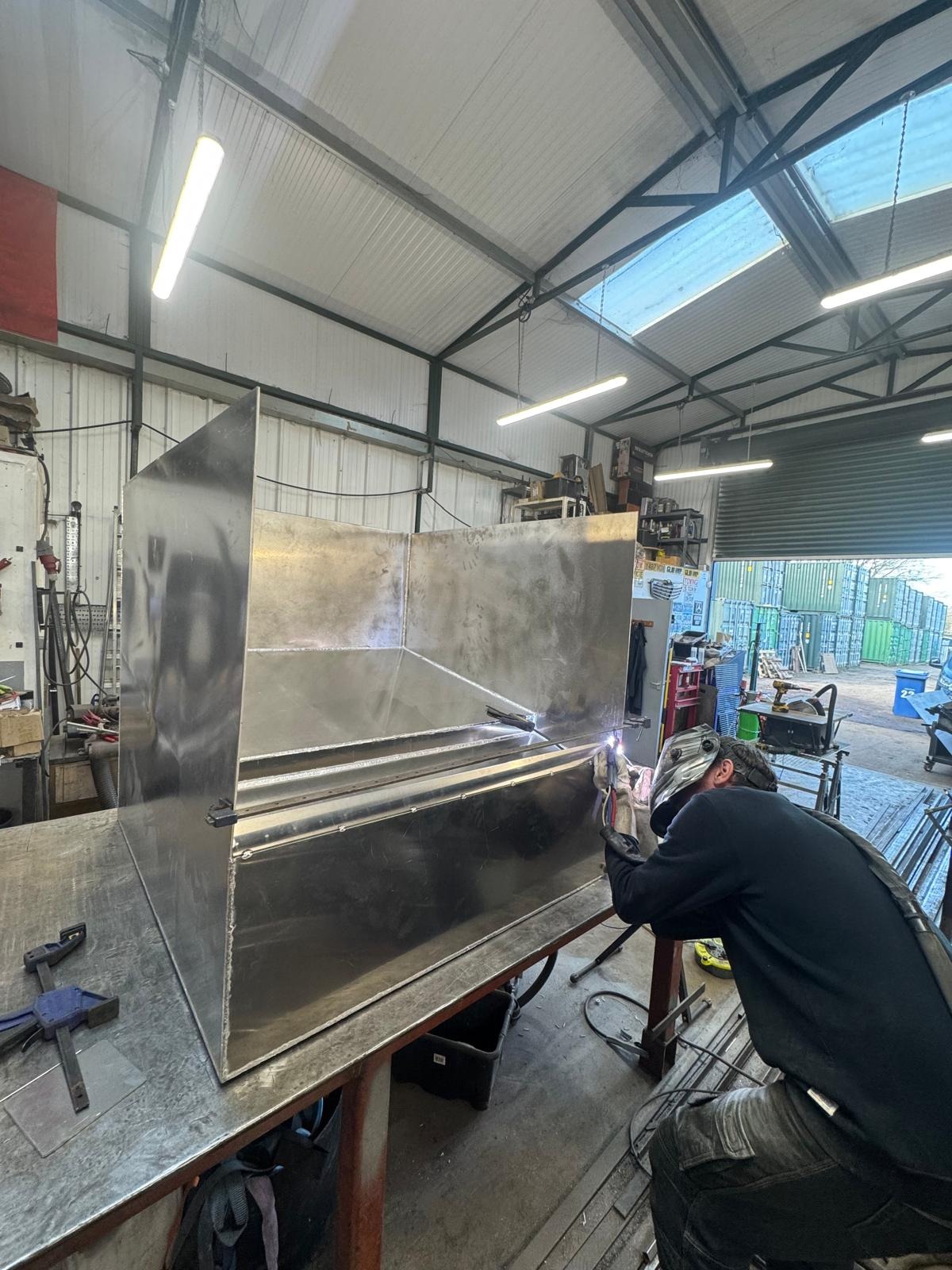 Spawacz w warsztacie łączy metalowe elementy dużej konstrukcji, używając spawarki. Widoczne iskry i wyposażenie warsztatu.