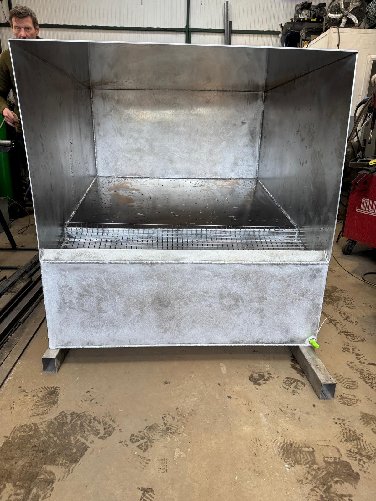 Surowa, metalowa konstrukcja przypominająca wannę spawalniczą na metalowych nogach, z kratką wewnątrz. Widoczne ślady pracy spawalniczej i niedoskonałości wykończenia.