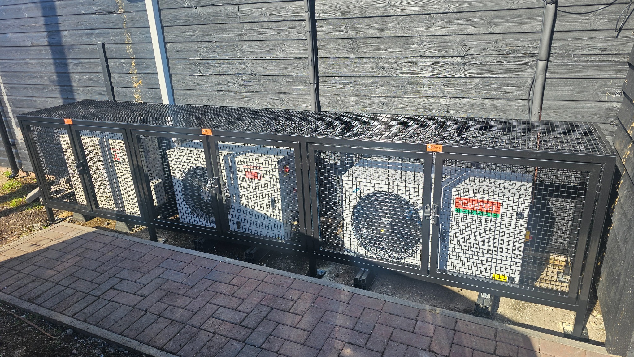 Czarna, metalowa konstrukcja z siatki, osłaniająca dwa białe agregaty klimatyzacyjne. Konstrukcja na niskich nóżkach, zamykana na zatrzaski. Tło: ciemna, drewniana ściana.