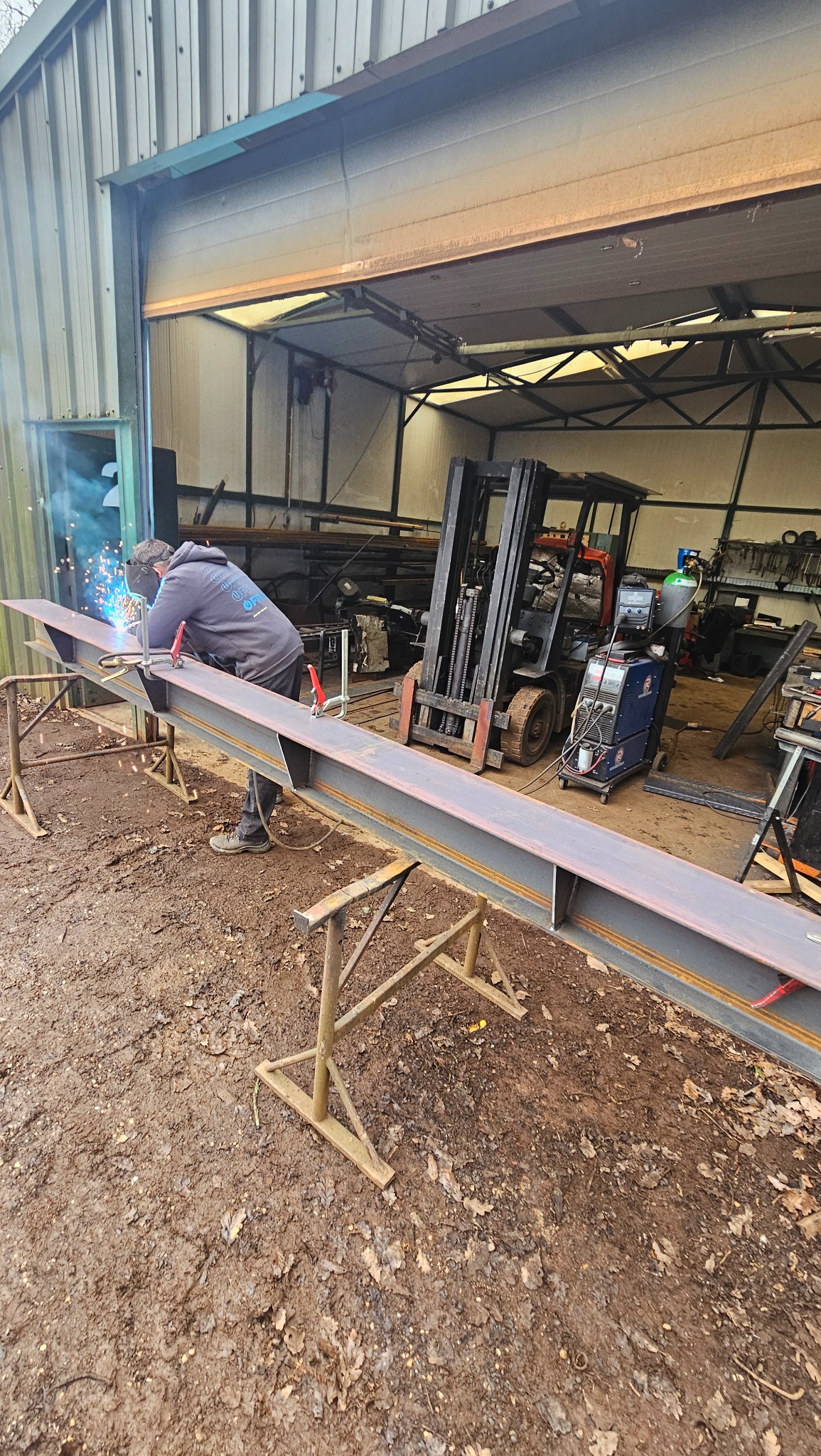 Spawanie metalowej belki H-kształtnej na kozłach w warsztacie, spawacz w masce, widoczny wózek widłowy i sprzęt spawalniczy. Podłoże ziemiste.