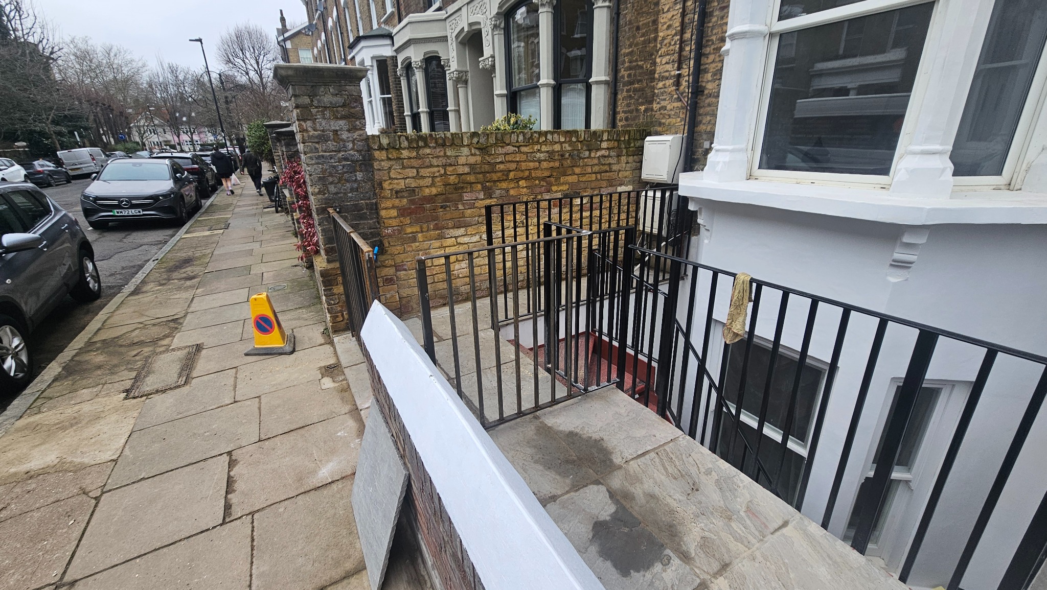 Metalowa barierka z furtką zabezpieczająca zejście do piwnicy w kamienicy. Widok z chodnika, w tle zaparkowane auta i przechodnie.