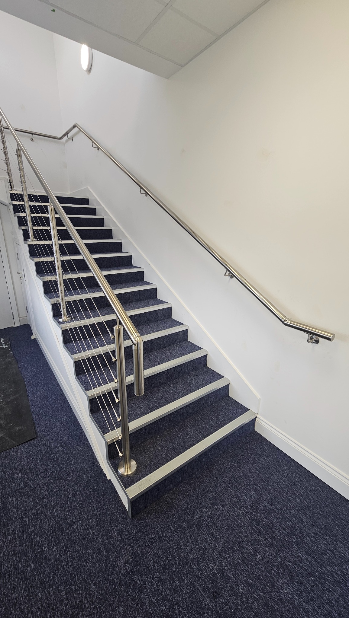 Nowoczesne schody wewnątrz budynku z granatową wykładziną i balustradą ze stali nierdzewnej z linkami, widziane z dołu. Minimalistyczny design.