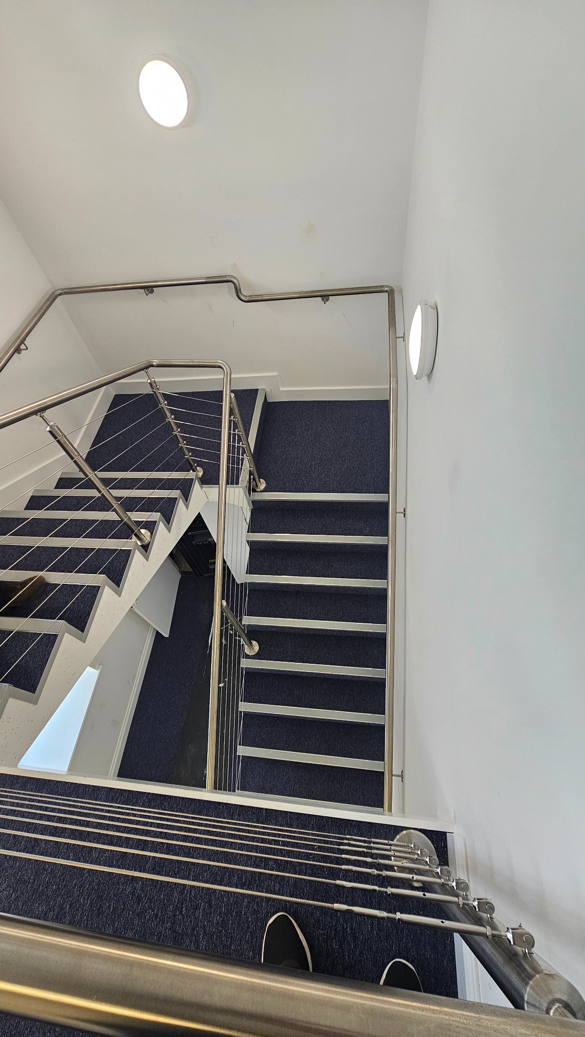 Widok z góry na schody z niebieską wykładziną i metalową balustradą z linkami. Ujęcie z perspektywy osoby stojącej na schodach. Widoczne oświetlenie sufitowe.