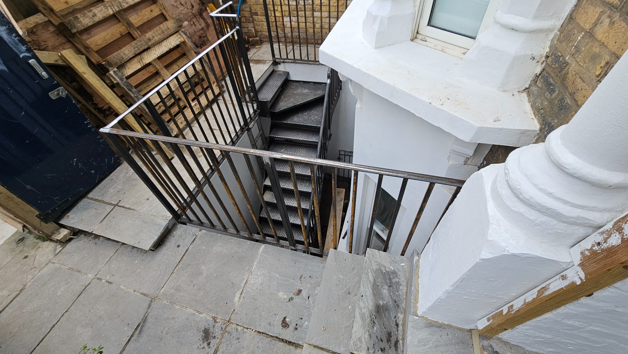 Metalowe schody ze stalową balustradą prowadzące w dół, umieszczone w przestrzeni przy budynku z elementami kamiennymi i drewnianymi paletami w tle.