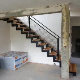 Surowe wnętrze z drewniano-metalowymi schodami z siatkową balustradą, widoczne materiały budowlane i niedokończone wykończenie ścian.