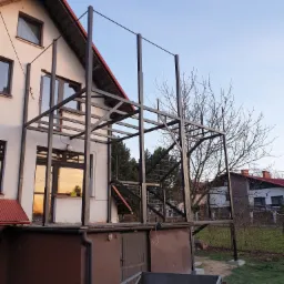 Metalowa konstrukcja tarasu na elewacji domu w trakcie budowy, widoczne schody i elementy nośne, w tle dom z czerwonym dachem i drzewo bez liści.