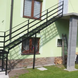 Zewnętrzne, czarne schody metalowe prowadzące na betonowy taras, z widoczną drabiną i stosem drewna obok, na tle jasnozielonego budynku.