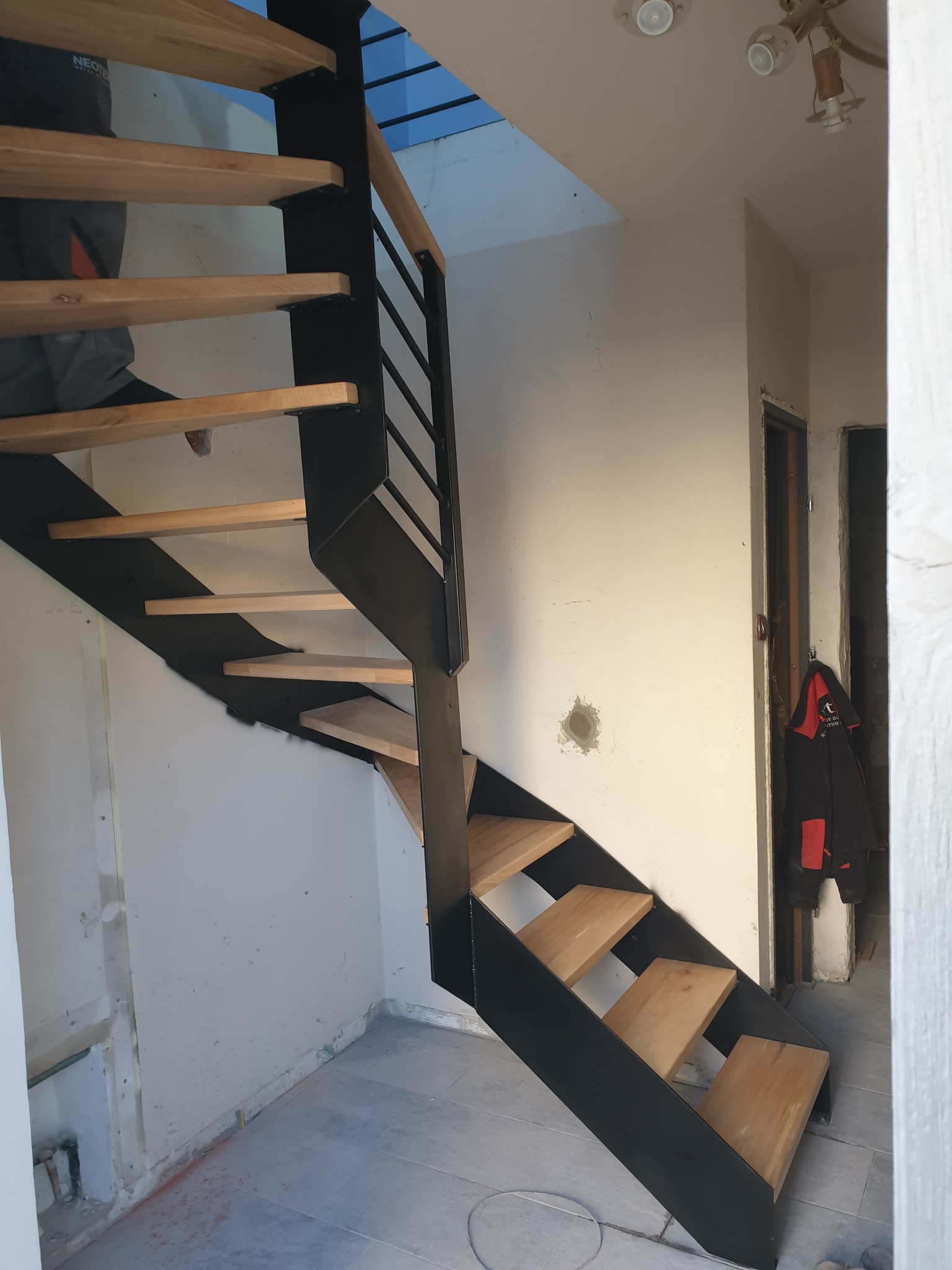 Nowoczesne schody z czarną metalową konstrukcją i drewnianymi stopniami w trakcie montażu, widoczne niedokończone wykończenia ścian.