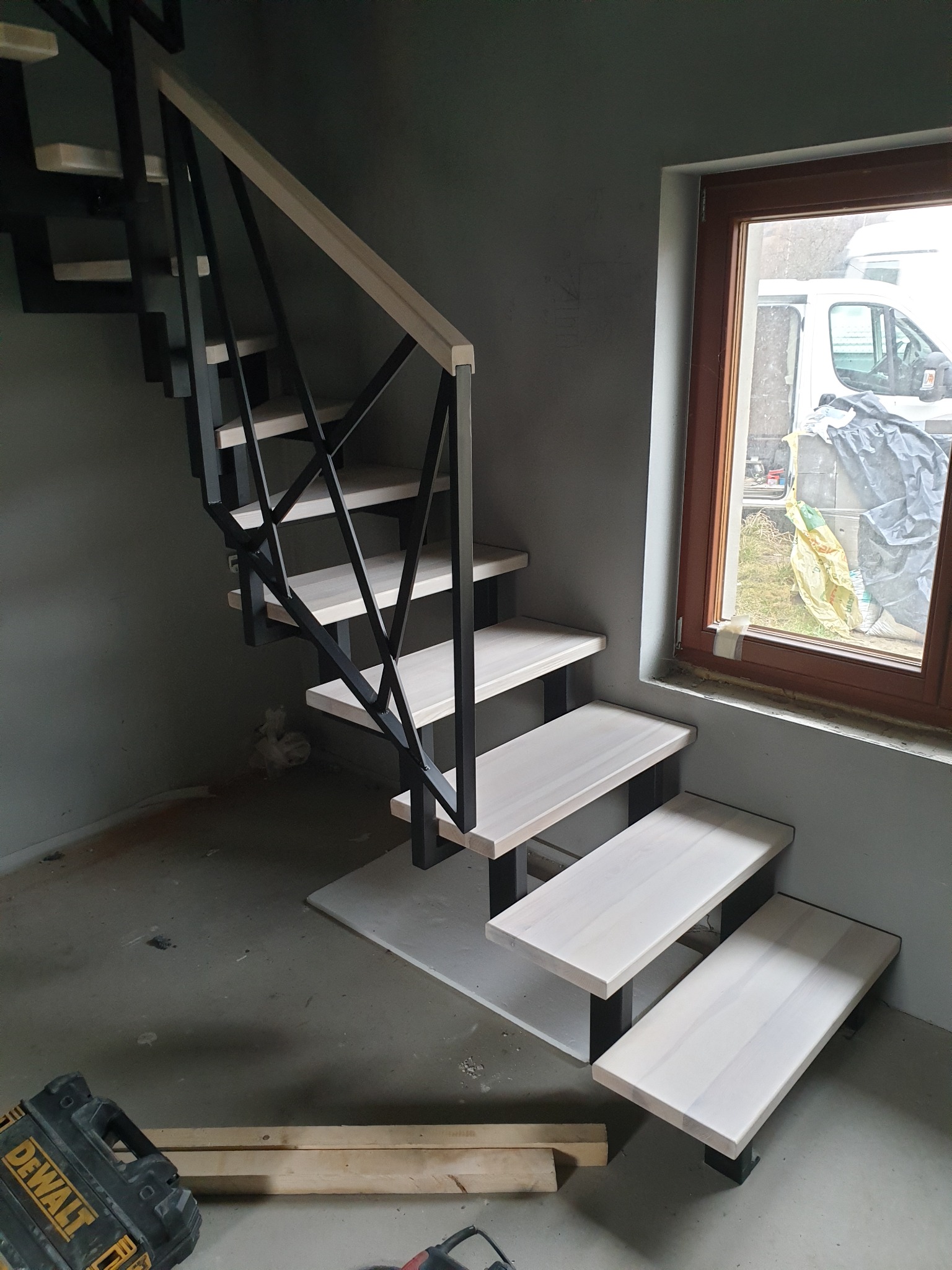 Nowoczesne schody metalowe z drewnianymi stopniami w surowym wnętrzu, widoczna skrzynka narzędziowa DeWalt i okno z widokiem na biały samochód dostawczy.