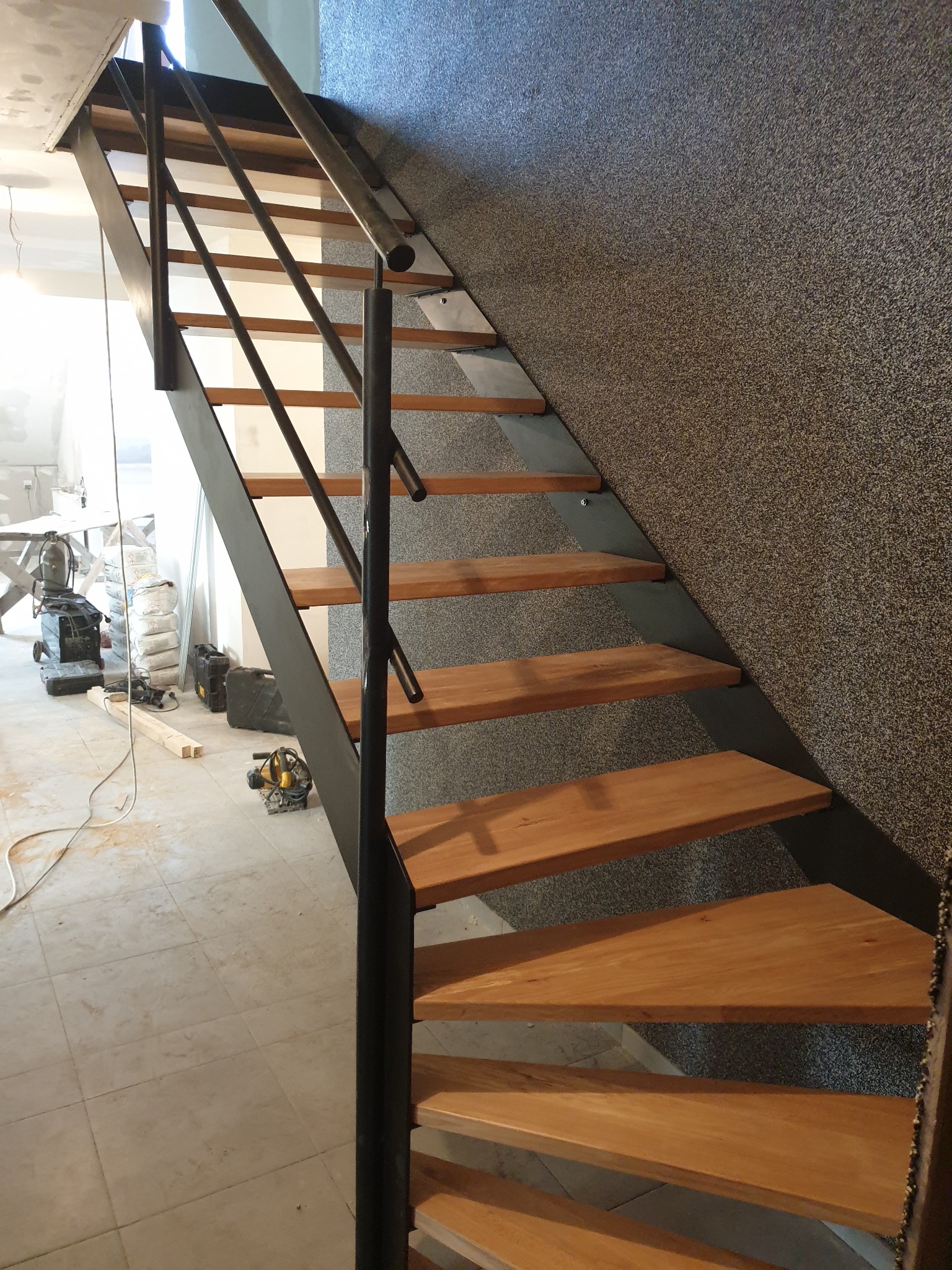 Nowoczesne schody z drewnianymi stopniami i metalową konstrukcją w trakcie montażu, widoczne narzędzia i worki z zaprawą w tle.