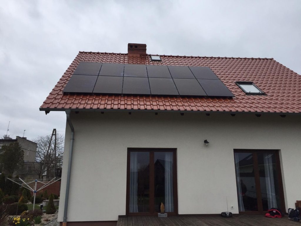 Czarny panel na dachu domu z czerwoną dachówką, widok z dołu na pochmurne niebo, dwa okna dachowe.