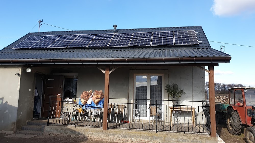 Dom jednorodzinny z panelami fotowoltaicznymi na dachu z blachodachówki, widoczny traktor z przyczepą drewna w tle.