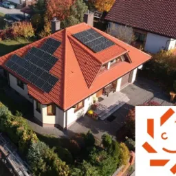 Widok z góry na dom jednorodzinny z czerwoną dachówką i panelami słonecznymi na dachu, otoczony zielenią, z widocznym logo firmy w prawym dolnym rogu.