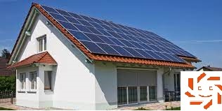 Biały dom jednorodzinny z ciemnoniebieskimi panelami fotowoltaicznymi na dachu, widok z niskiej perspektywy, z roletami częściowo zasłoniętymi w oknach na parterze i logo w prawym dolnym rogu.