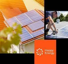 Montaż paneli słonecznych na dachu domu w Otwocku, widok z dołu, z fragmentem zieleni na pierwszym planie, obok wklejone zdjęcie montera z wiertarką oraz logo firmy Happy Energy.