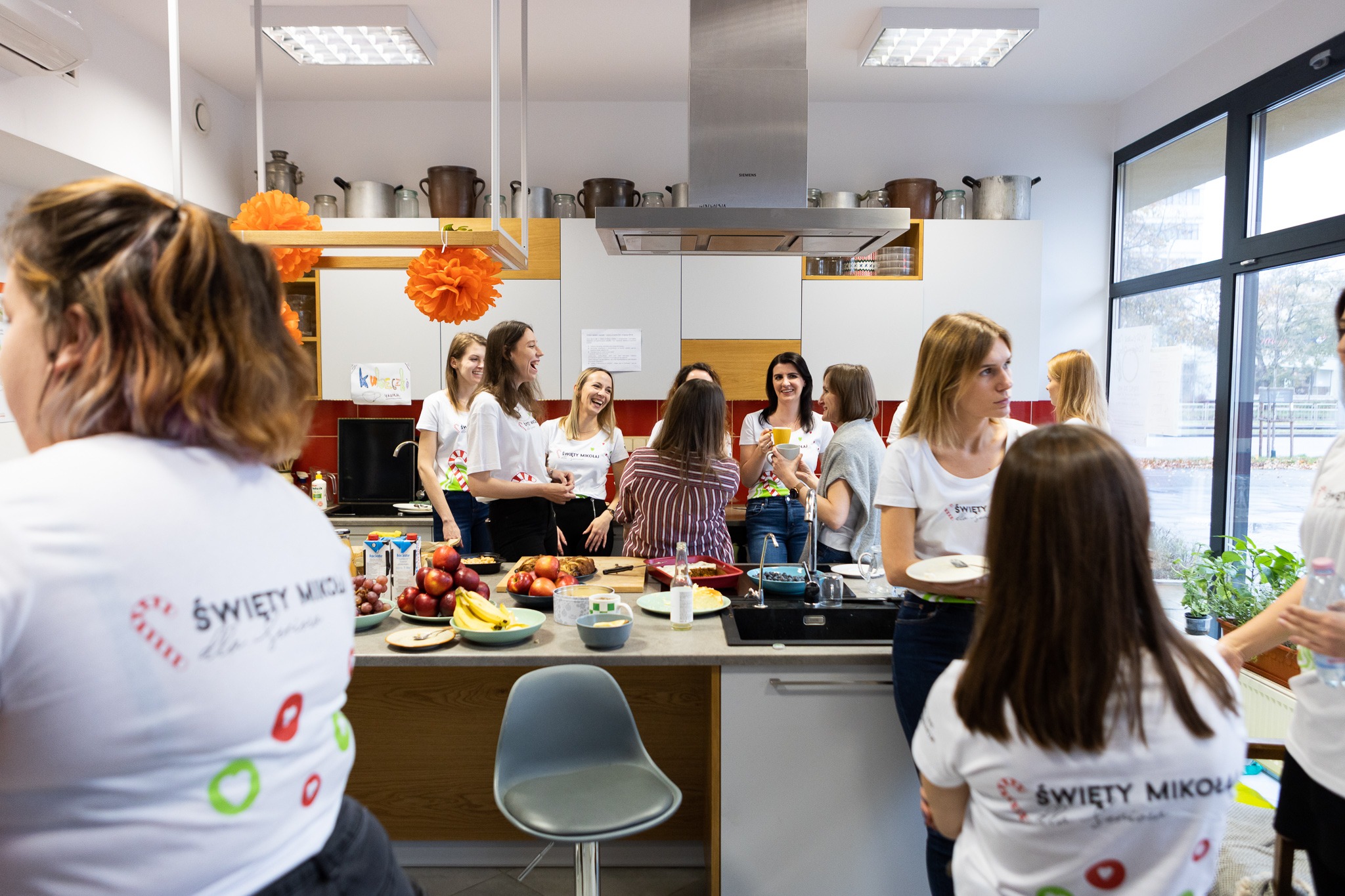 Grupa kobiet w kuchni, ubrane w białe koszulki z napisem 'Święty Mikołaj dla seniora', przygotowuje posiłek. Na blacie owoce, ciasta i napoje. Pomarańczowe dekoracje wiszą pod sufitem.