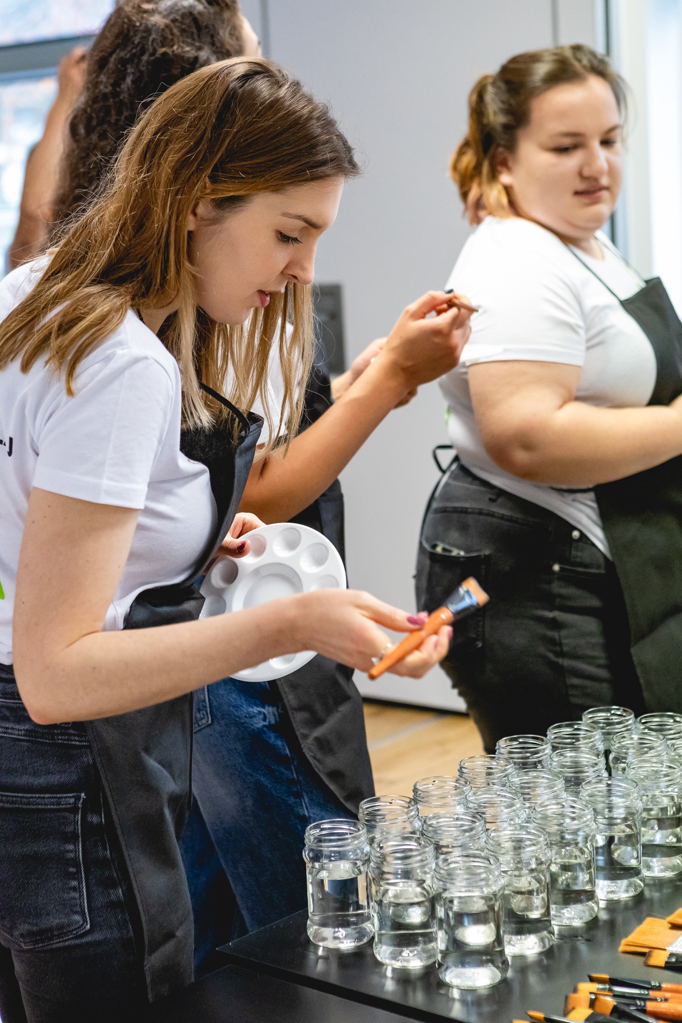 Warsztaty malowania na szkle: uczestniczka miesza farby na palecie, obok rzędy słoików z wodą i pędzle.